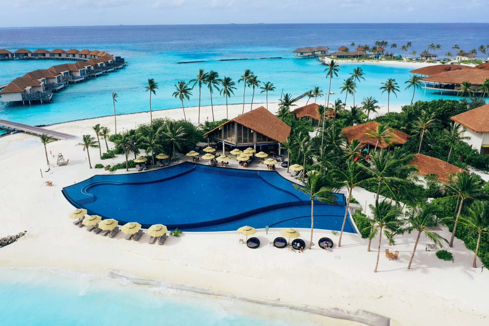 The main pool from above, free-form, with loungers, the beach and the overwater villas behind.