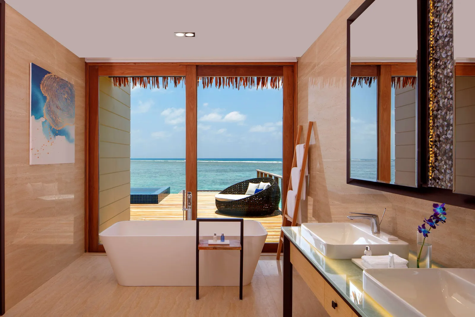 An overwater villa bathroom, with a freestanding bath, twin basins and the deck over the sea beyond the glass.