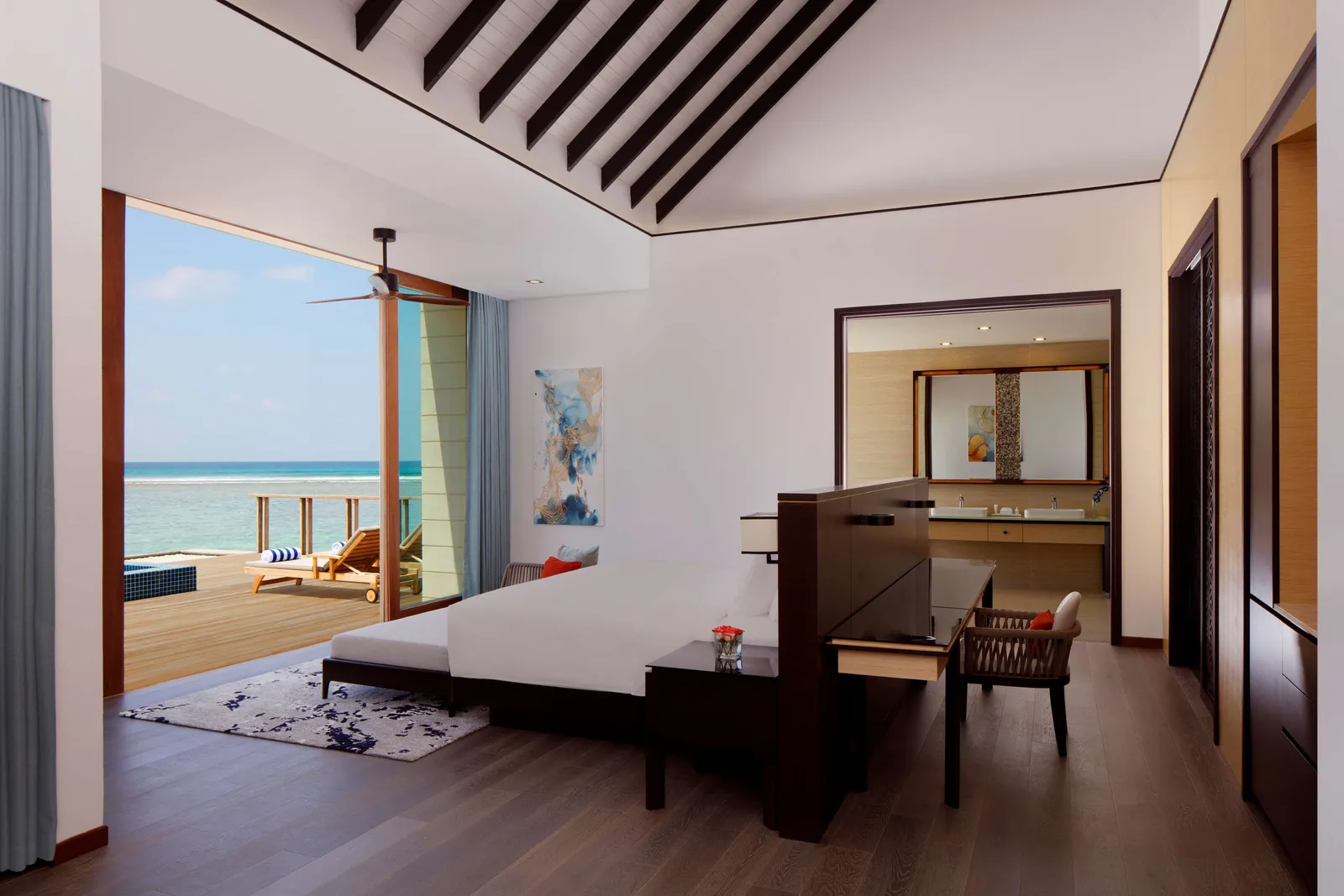 An overwater villa bedroom opening on to the deck, its loungers and the sea beyond the glass.