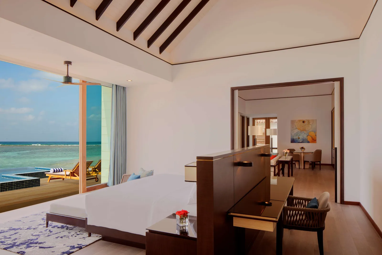 A bedroom of a 3-bedroom overwater villa, with a white bed, a desk and the pool beyond the French window.