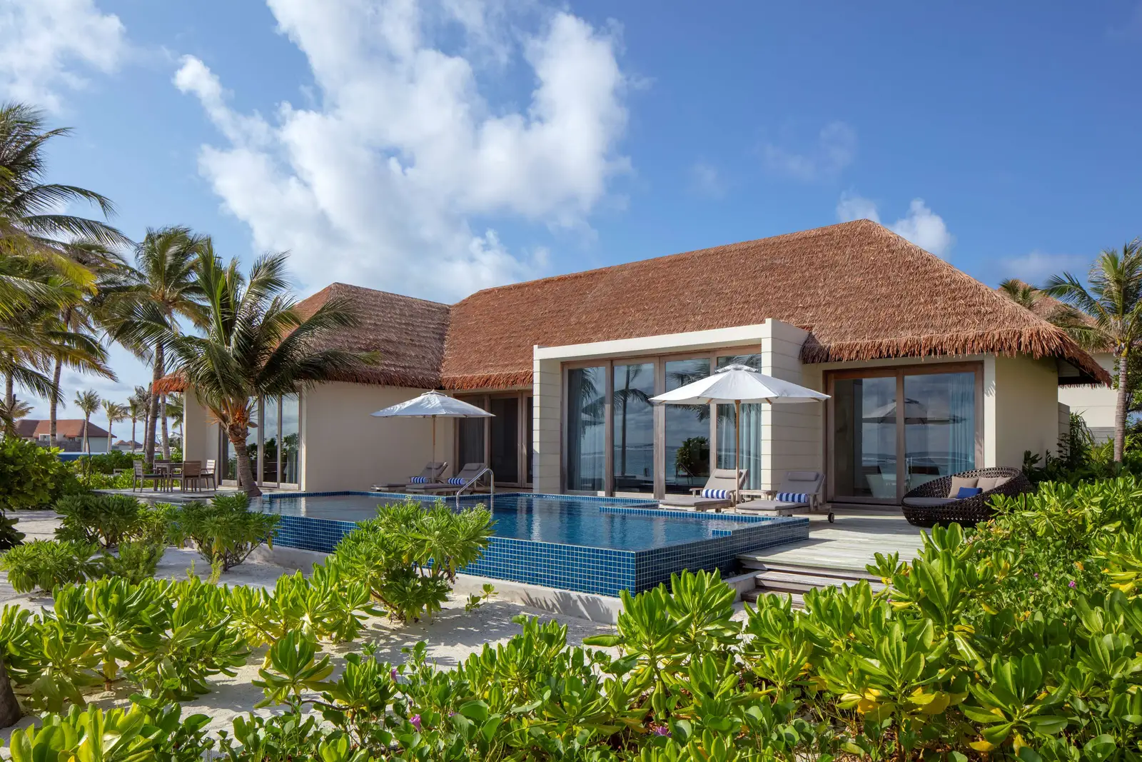 A 2-bedroom beach villa, its long thatched roof, parasols and pool among the greenery.