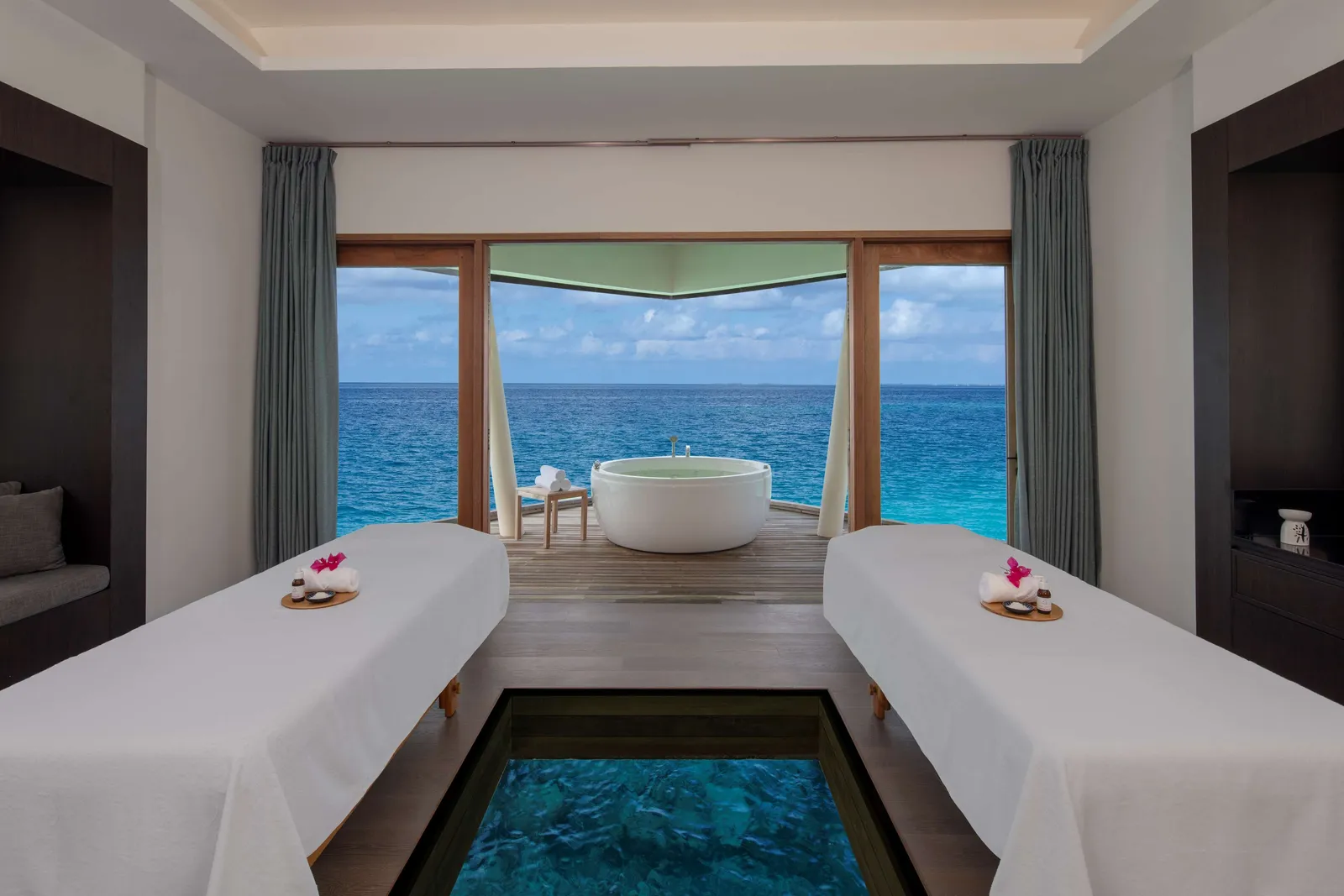 A Blu Spa room with 2 massage beds on a glass floor and a bath facing the sea.