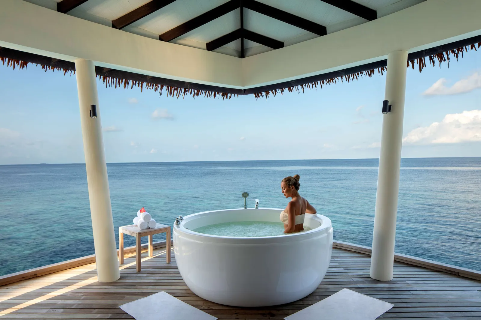 A woman in a round bath on the spa’s deck, under the thatch, facing the open sea.
