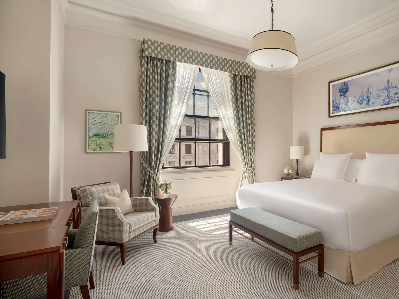 A Deluxe room: a white bed, an upholstered bench, a check armchair and 2 tall windows