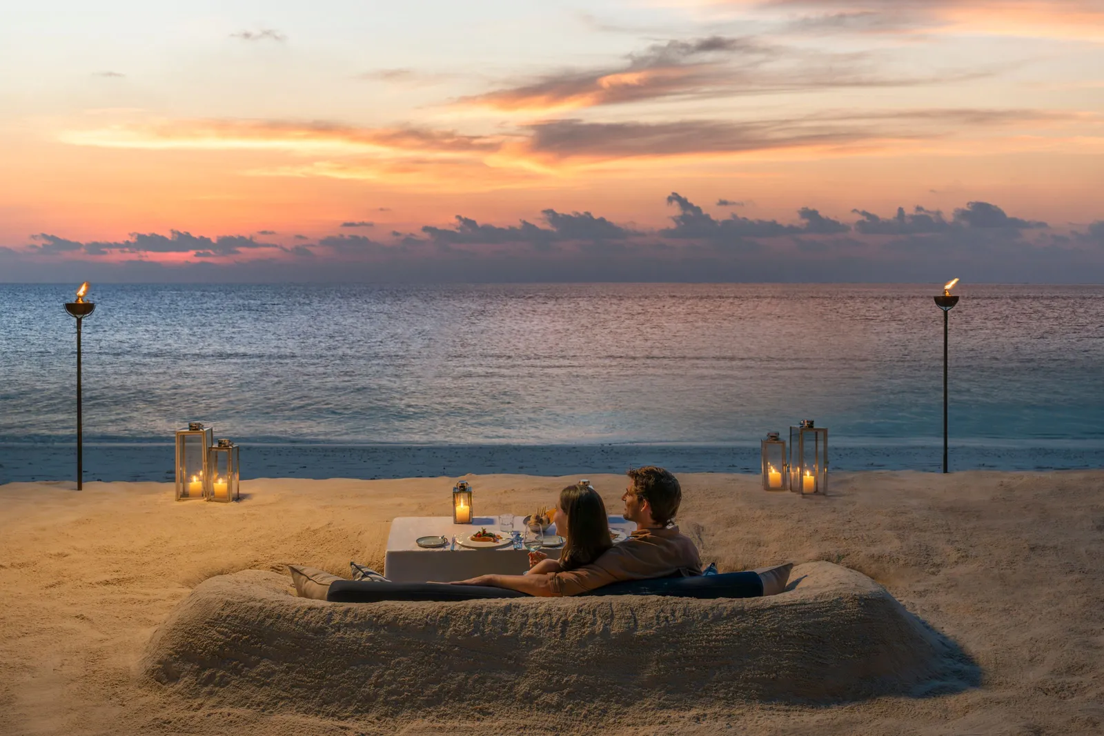 A couple dining at sunset at a table sunk into the sand among lanterns