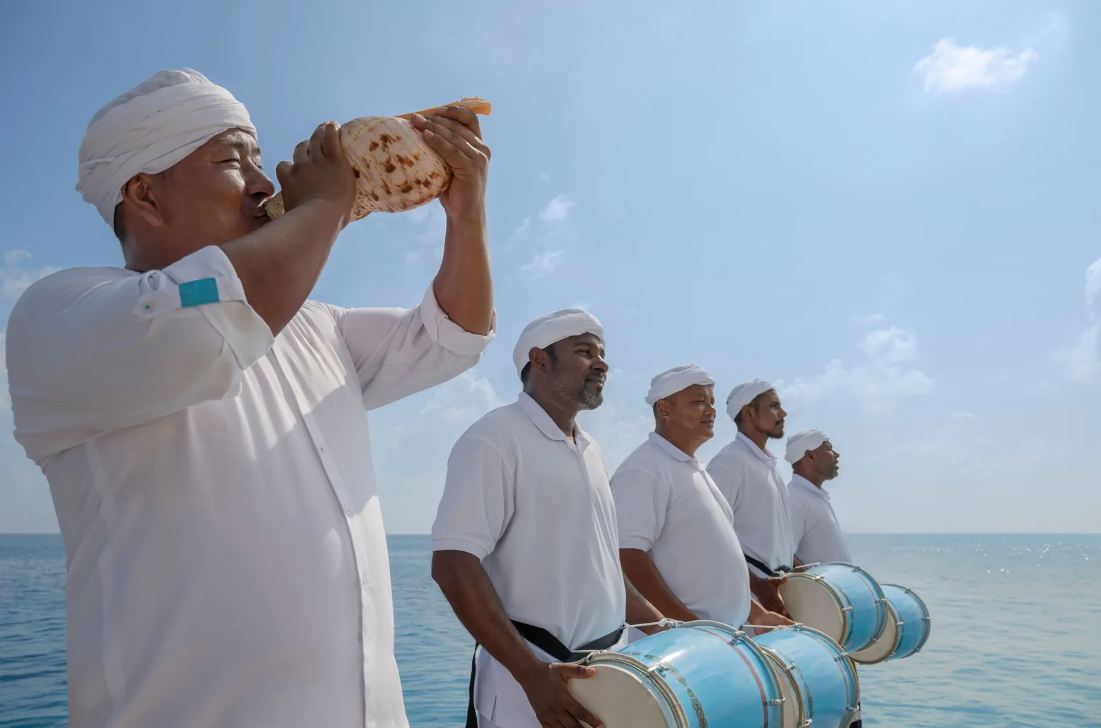 4 men in white play the bodu beru drums and blow a conch shell to welcome guests