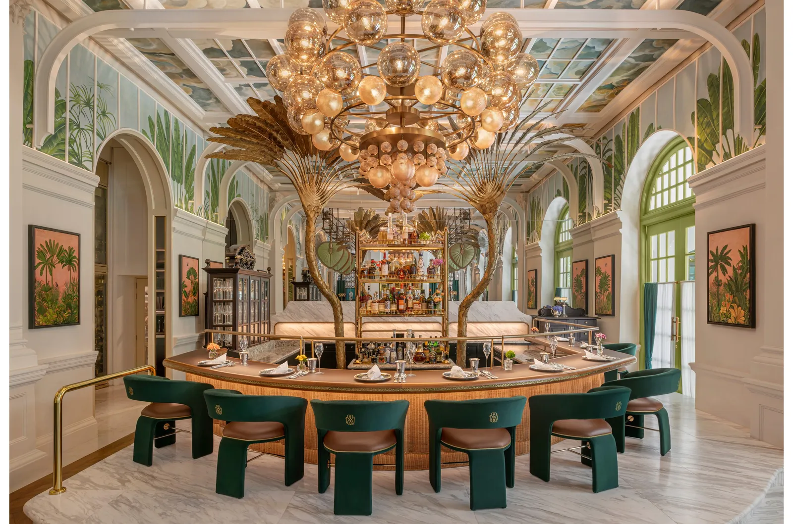 The dining room of 1887 by André: a circular counter with green armchairs under a gilded chandelier and tall metal palm fronds.