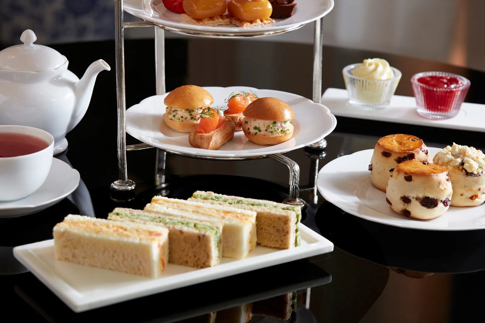 Afternoon tea close up: finger sandwiches, filled rolls, fruit scones with cream and jam, and a white teapot.