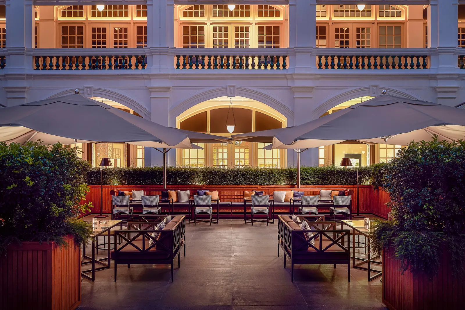 Raffles Courtyard in the evening: outdoor tables and sofas under parasols, in front of the arcade’s lit façade.