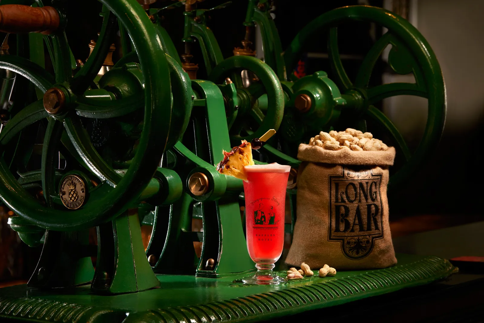 A Singapore Sling and the Long Bar’s sack of peanuts in front of the green cast-iron wheels of the machine on the counter.