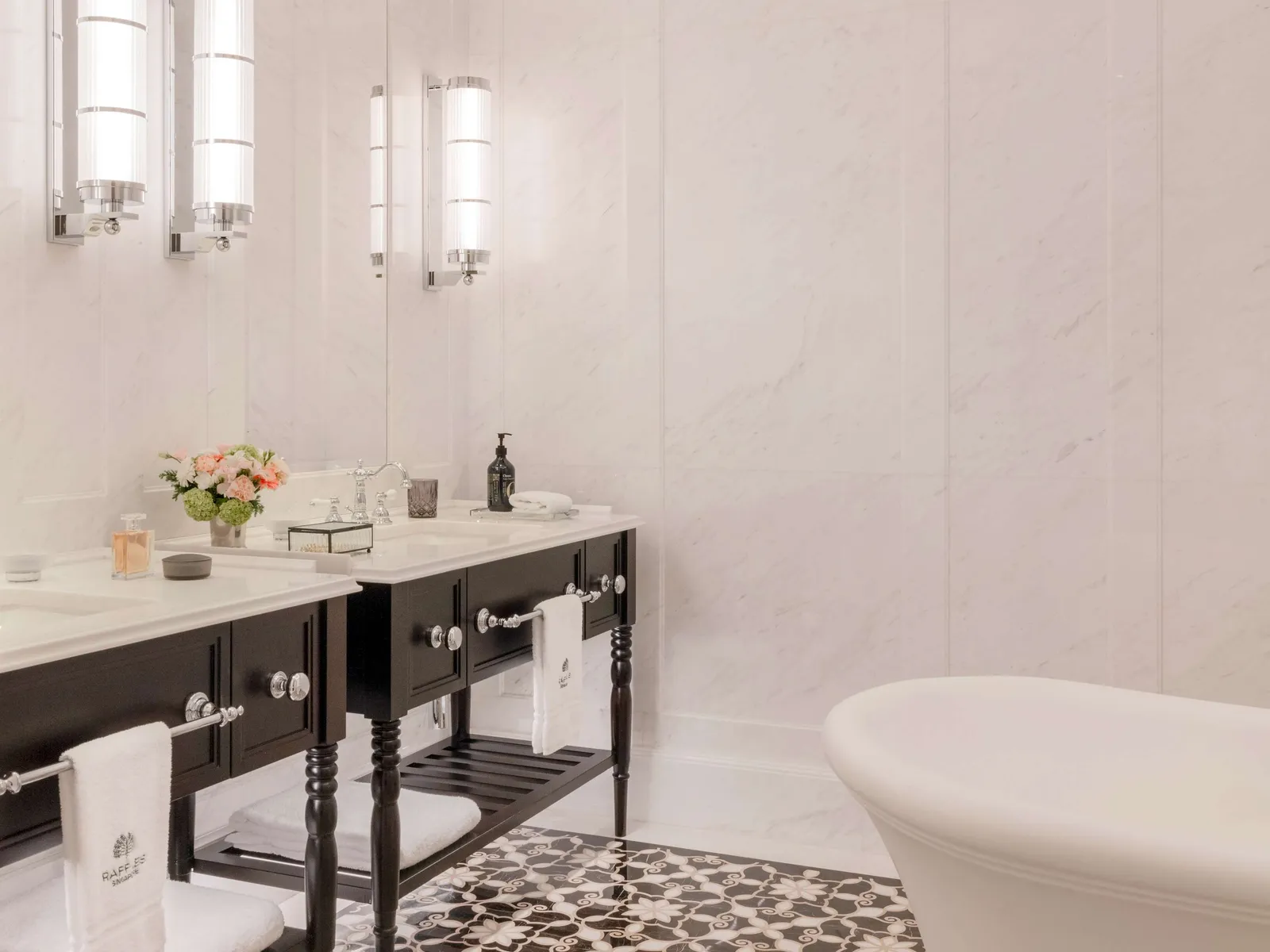 A suite’s bathroom: twin basins on a dark stand, white marble walls, the bath and a mosaic floor.