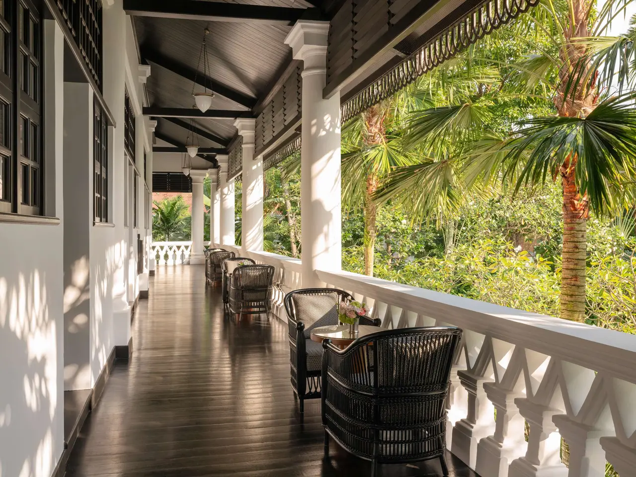 The dark-wood verandah outside the Palm Court Suites: rattan armchairs, the white balustrade and the garden’s palms.