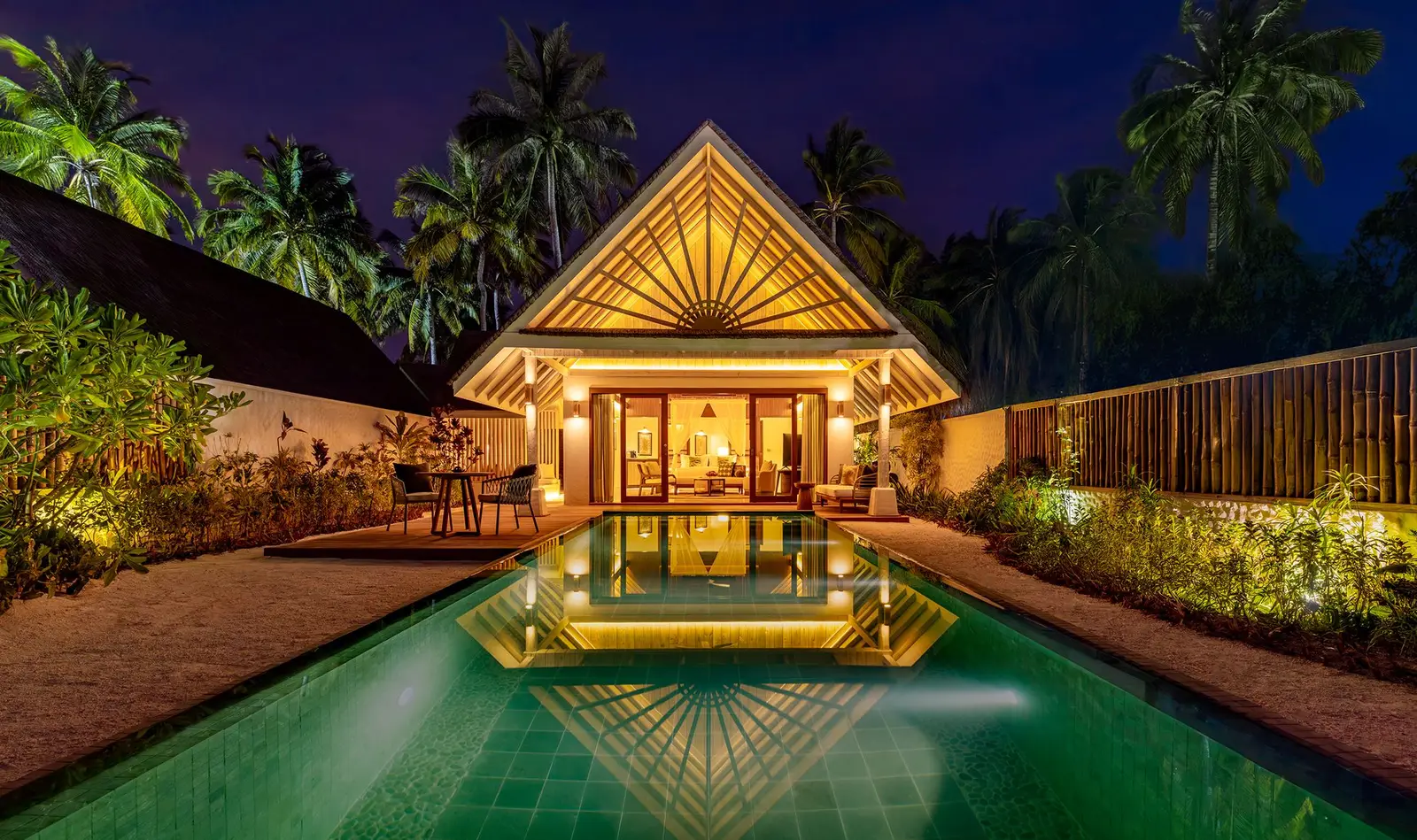 A beach villa at night, its sunburst gable lit up and mirrored in the long private pool among the palms.