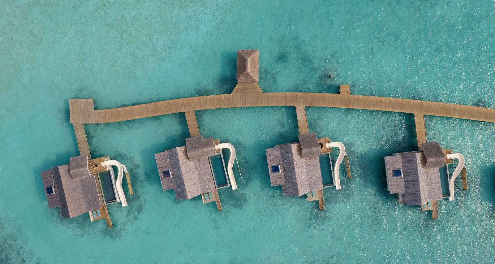 The water villas seen from above along the jetty, each with its timber roof and a white slide curving down into the lagoon.