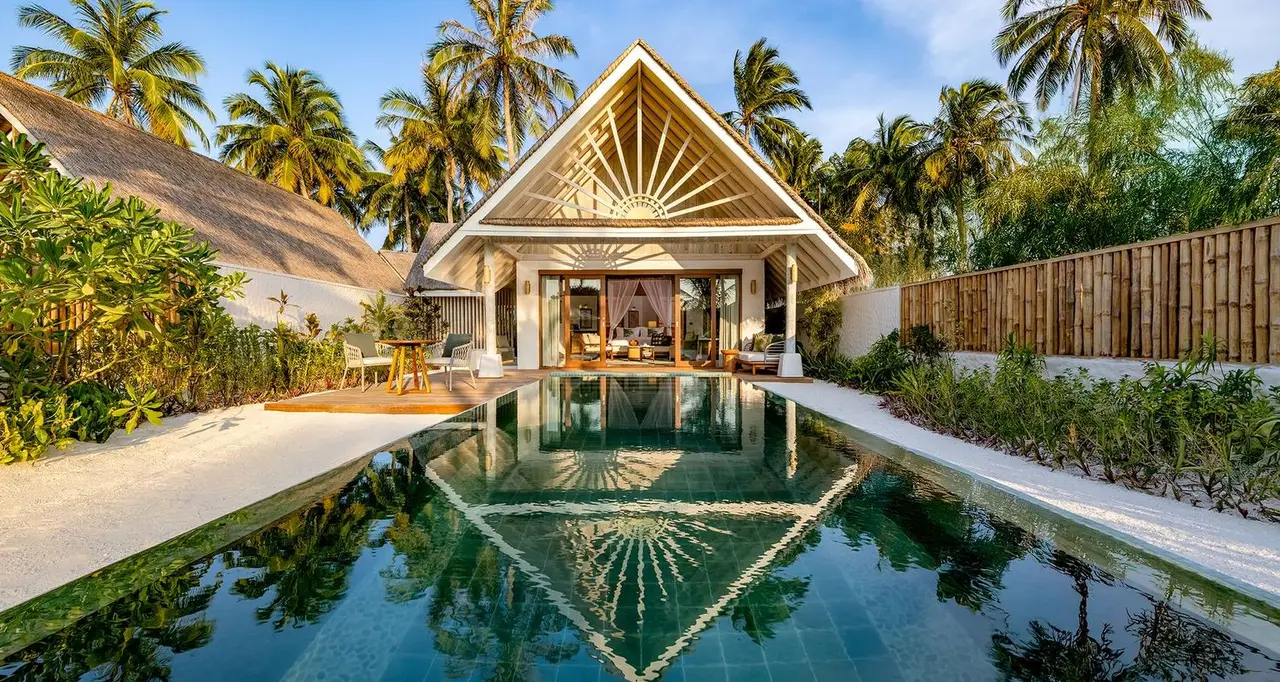 The long pool of a Beach Pool Villa in front of the white villa with its sunburst gable, among palms and shrubs.