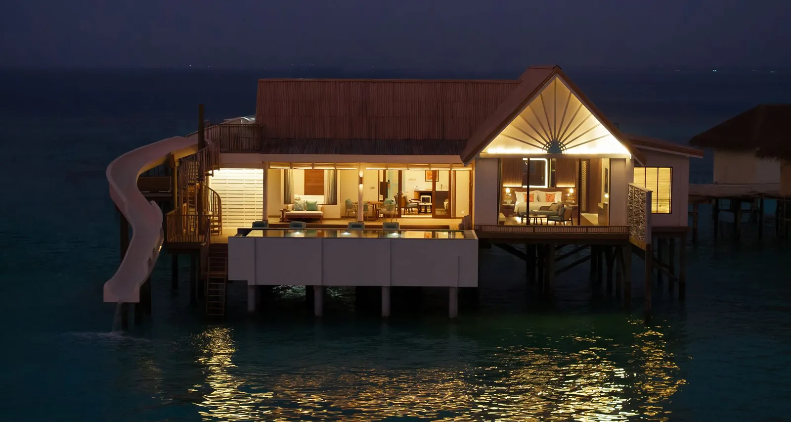 The Sunset Family Water Pool Villa at night, raised over the lagoon, with lit windows and the slide at its side.