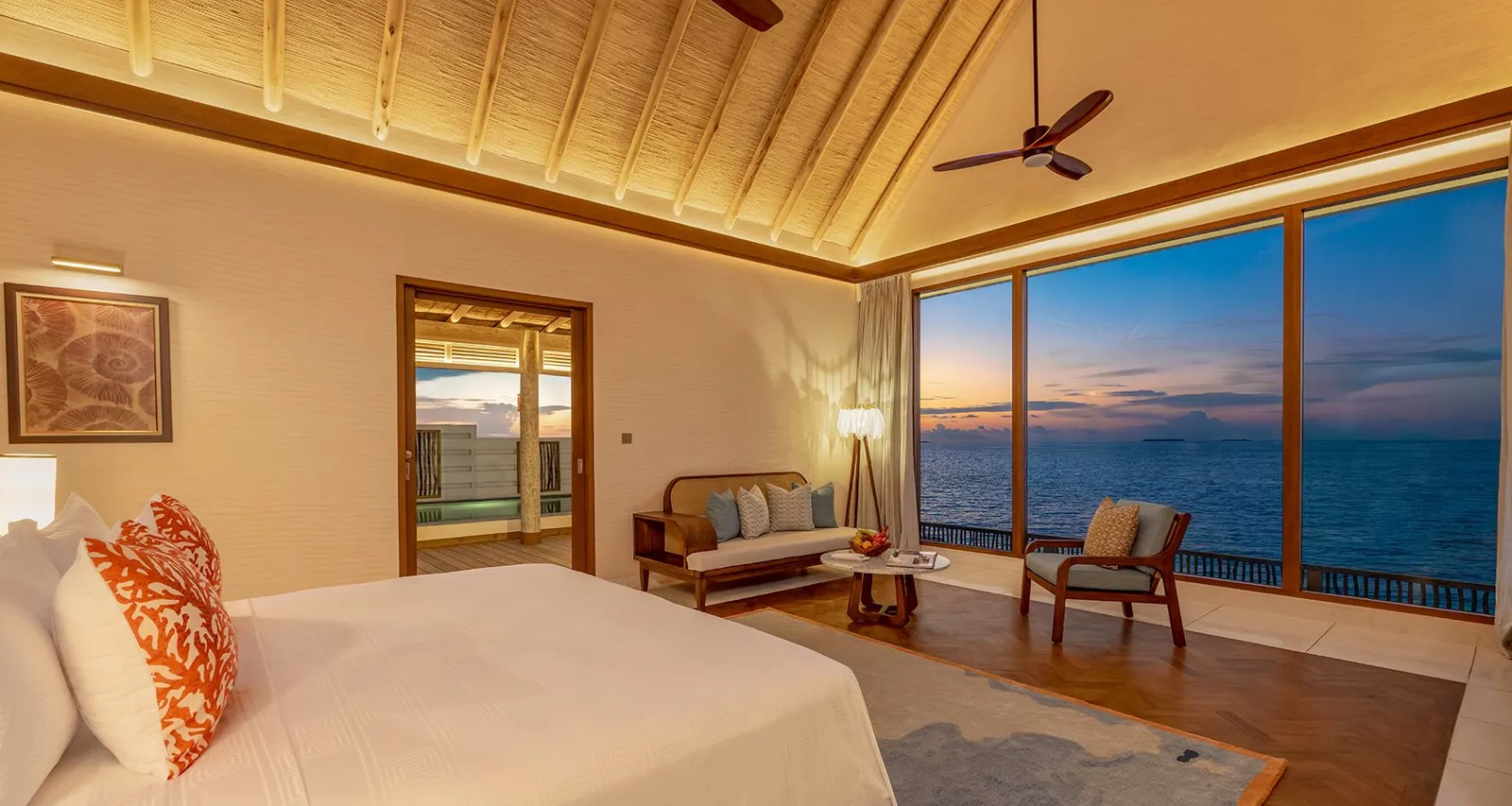 A Sunset Water Pool Villa’s bedroom at sunset, with the bed, armchairs and a wall of glass onto the sea.