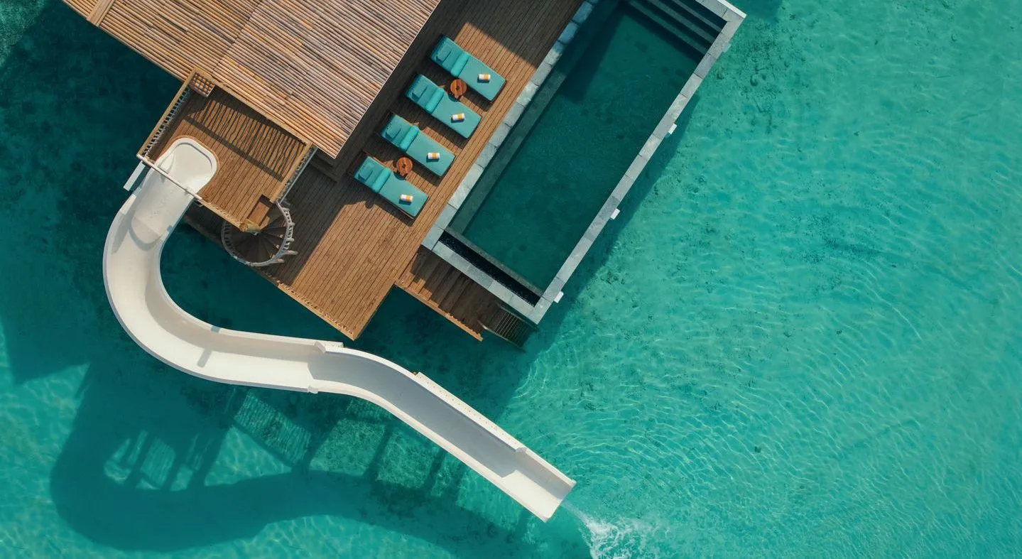 A water villa from above: the deck with its loungers, the infinity pool and the slide ending in the lagoon.
