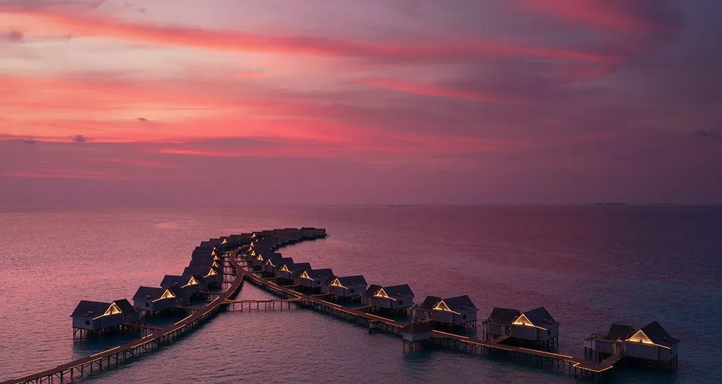The jetty of water villas at sunset, its lights on under a pink sky.