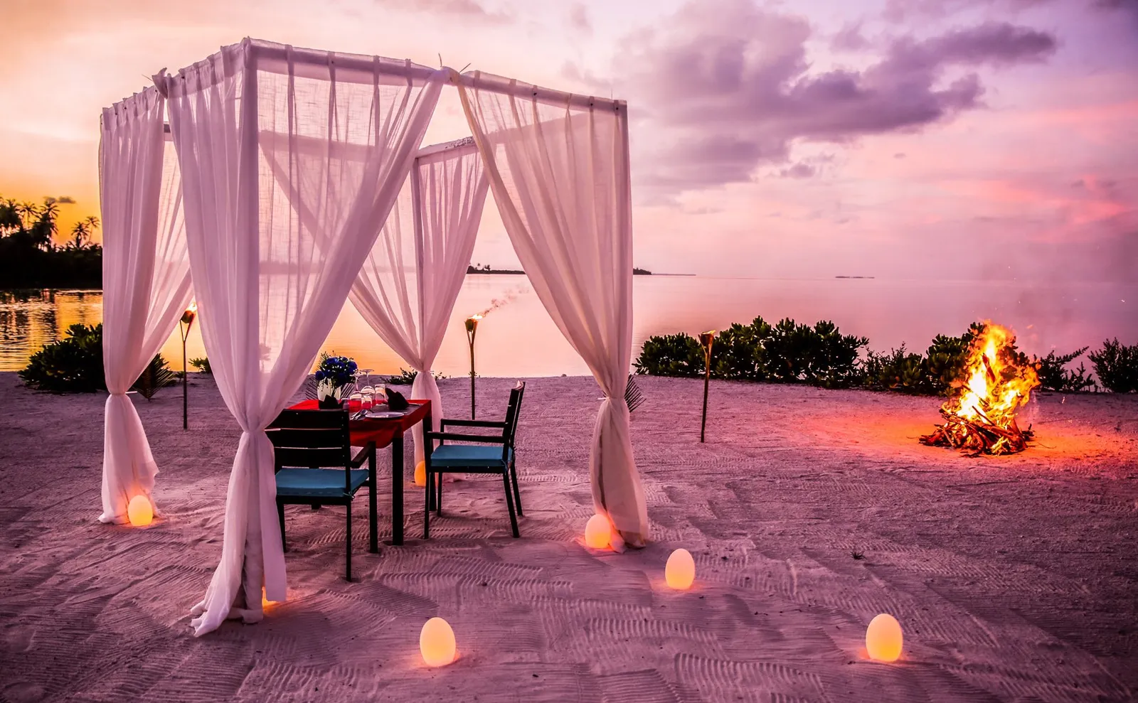 A beach dinner at sunset: the white-curtained pavilion, the table for 2, lanterns and a bonfire.