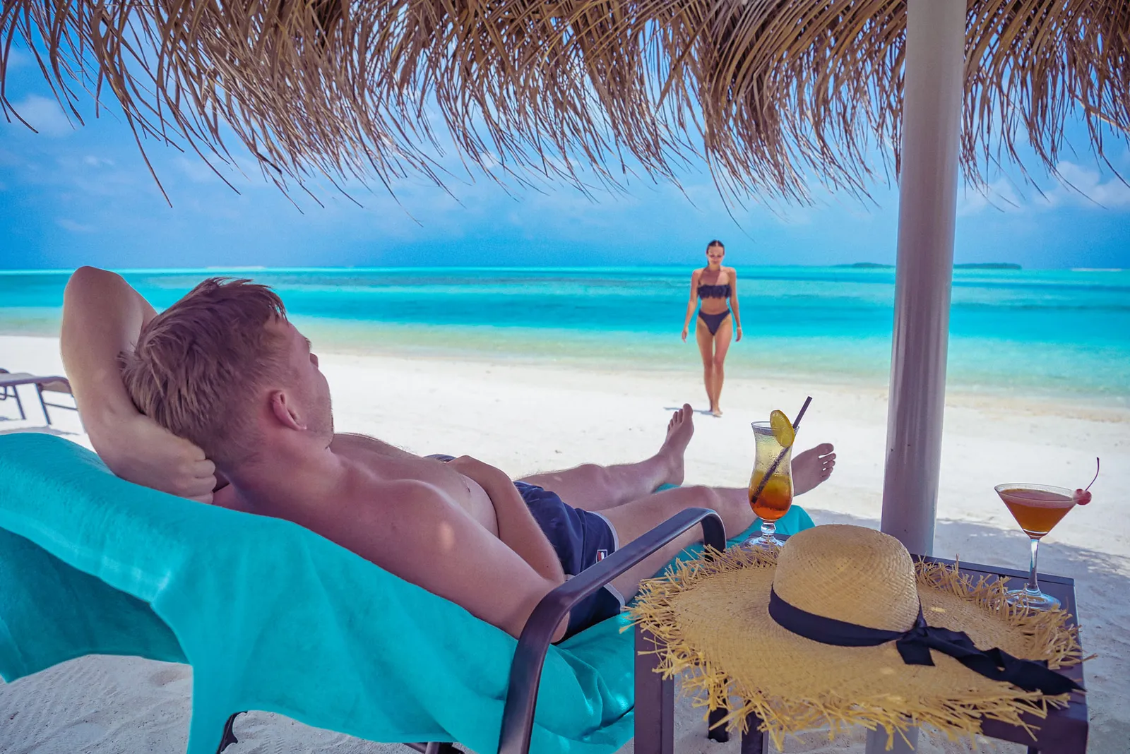 A guest on a lounger in the shade at Dhirun’Bar, a cocktail and a straw hat beside him, the turquoise sea ahead.