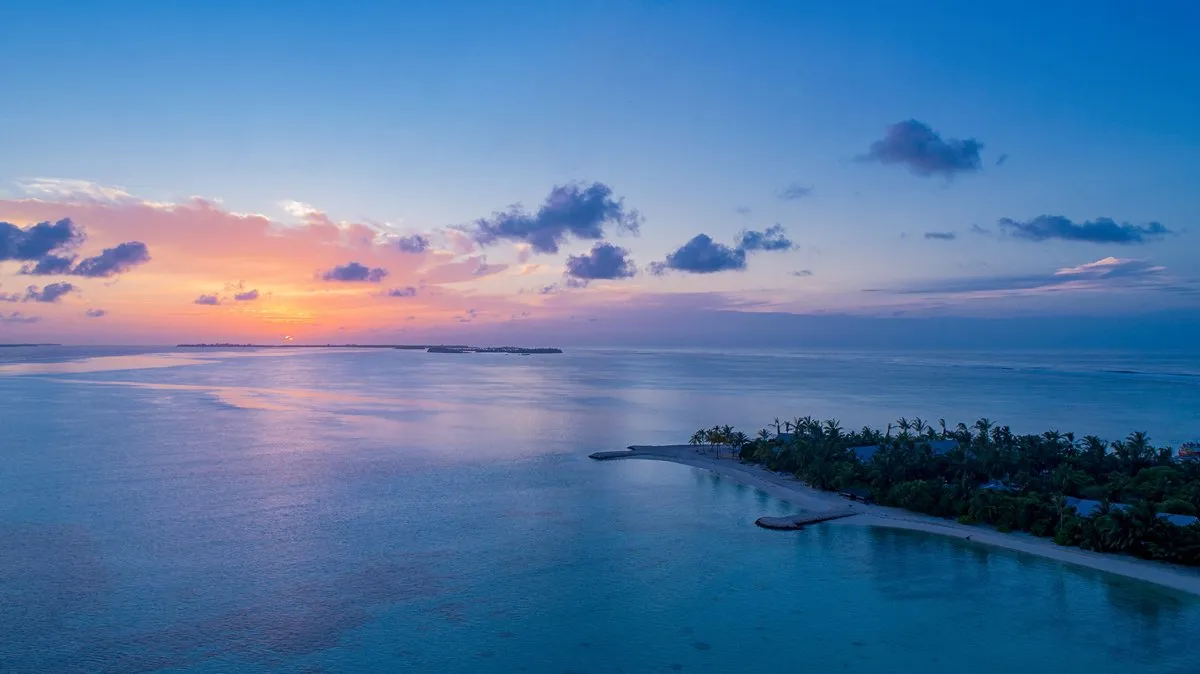 Dawn over the ocean from above, with the palm-covered tip of the island and the shallow lagoon around it.