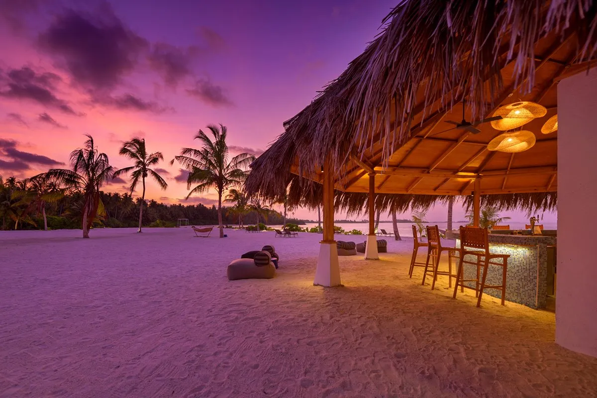 Ko’Bar at sunset: the lit thatched roof, the stools at the counter and the palms against a violet sky.