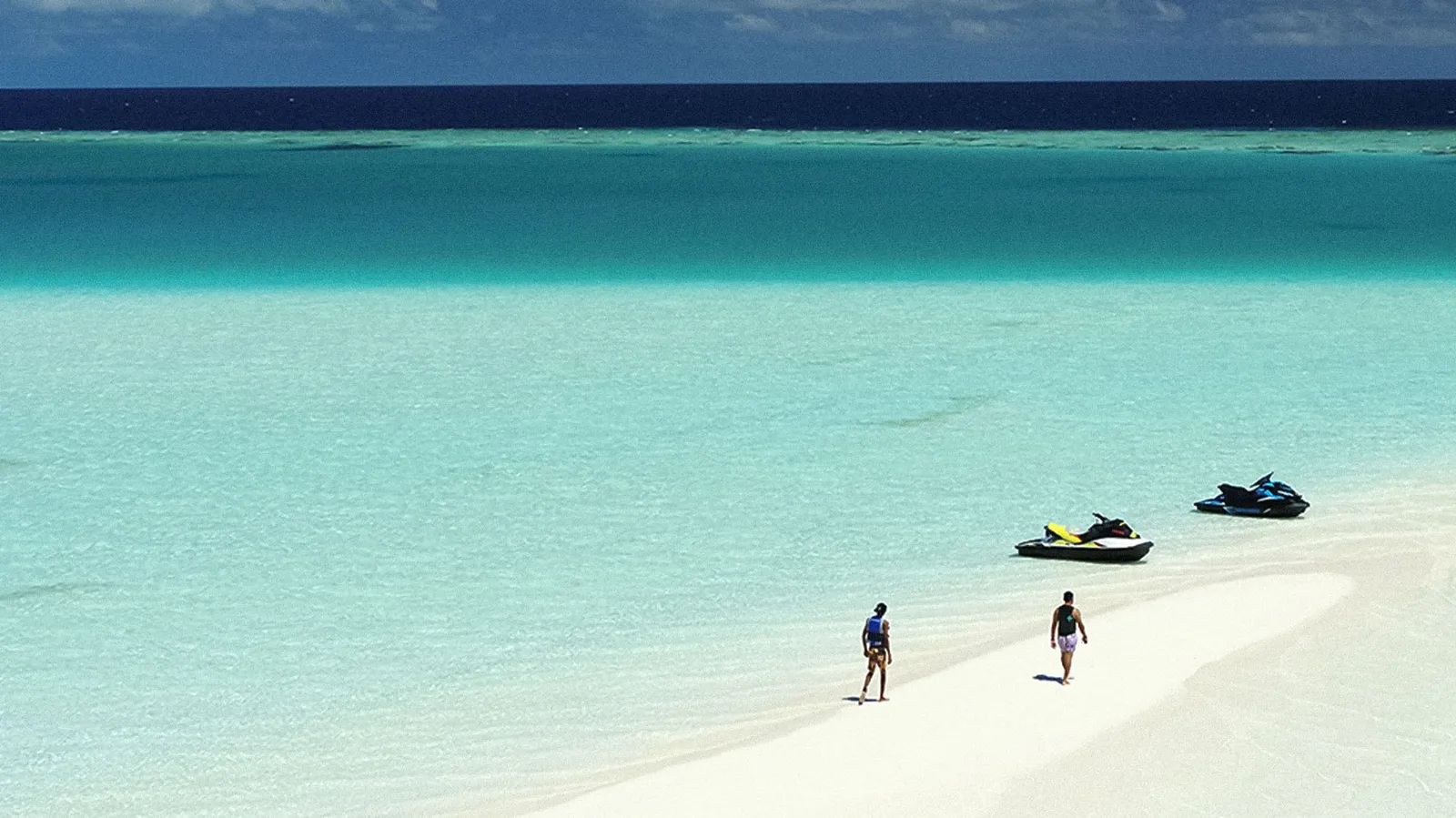 2 people walking on a sandbank in the lagoon, 2 jet skis pulled up at the water’s edge.