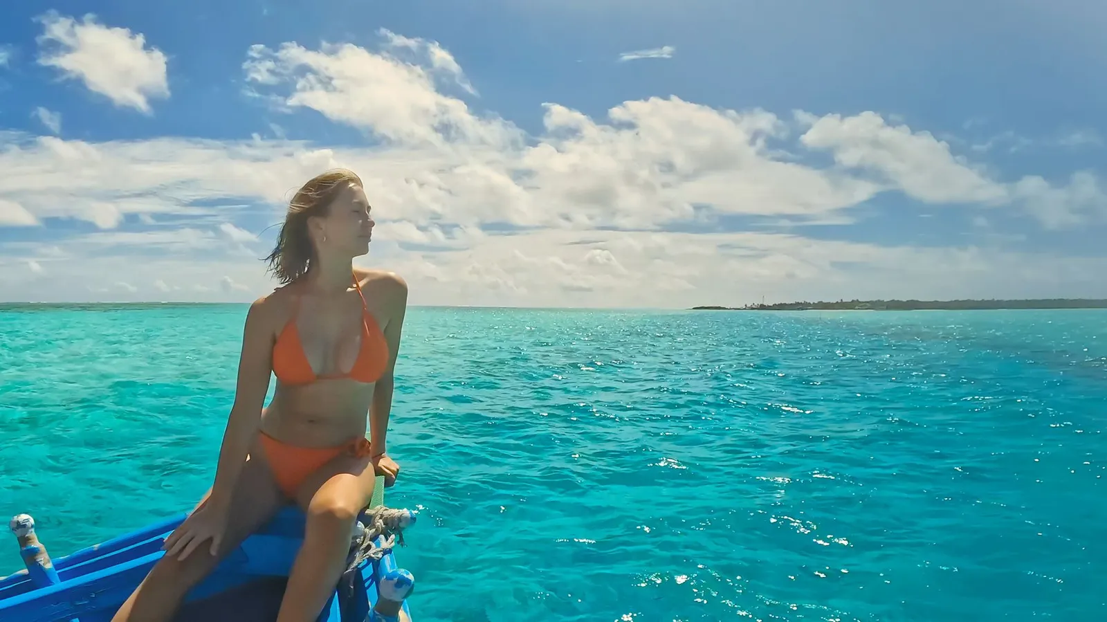 A guest sitting at the bow of a blue dhoni, on open turquoise water with a low island on the horizon.