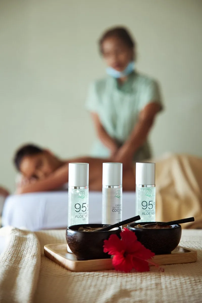 A Rahaa Spa room: a therapist massages a guest on the couch, oils and a hibiscus in the foreground.