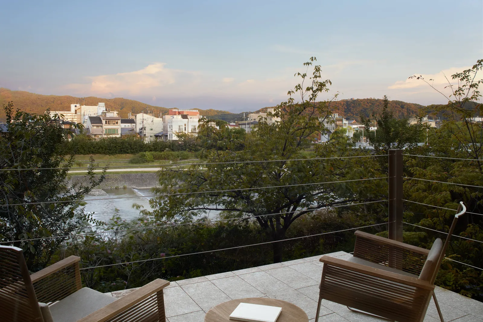 2 armchairs on a stone terrace above the Kamo River, among the maples, with the houses of the far bank and the Higashiyama hills.