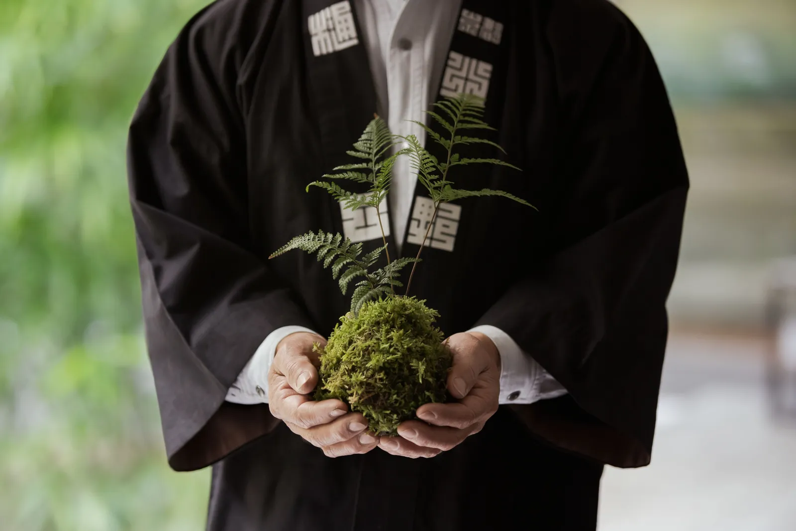 A gardener in a traditional jacket holding a kokedama, a ball of moss with a fern growing from it.