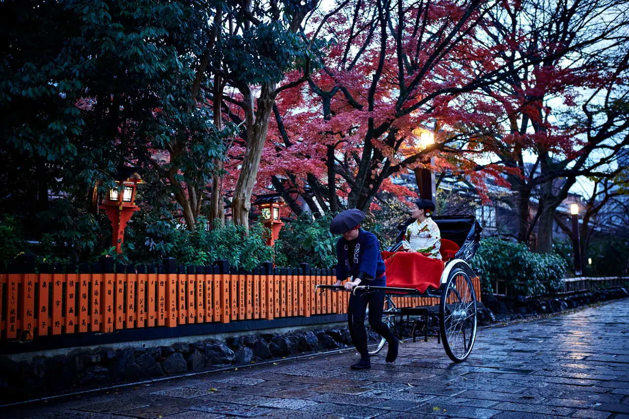 A rickshaw pulled along a wet stone street in Gion, under red autumn maples and lanterns.