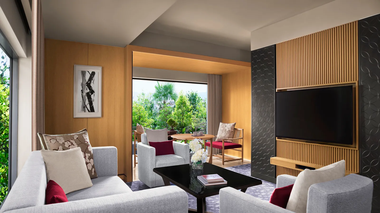 The Corner Suite KITA living room: grey sofa and armchairs, the television on a timber wall and the garden beyond the window.