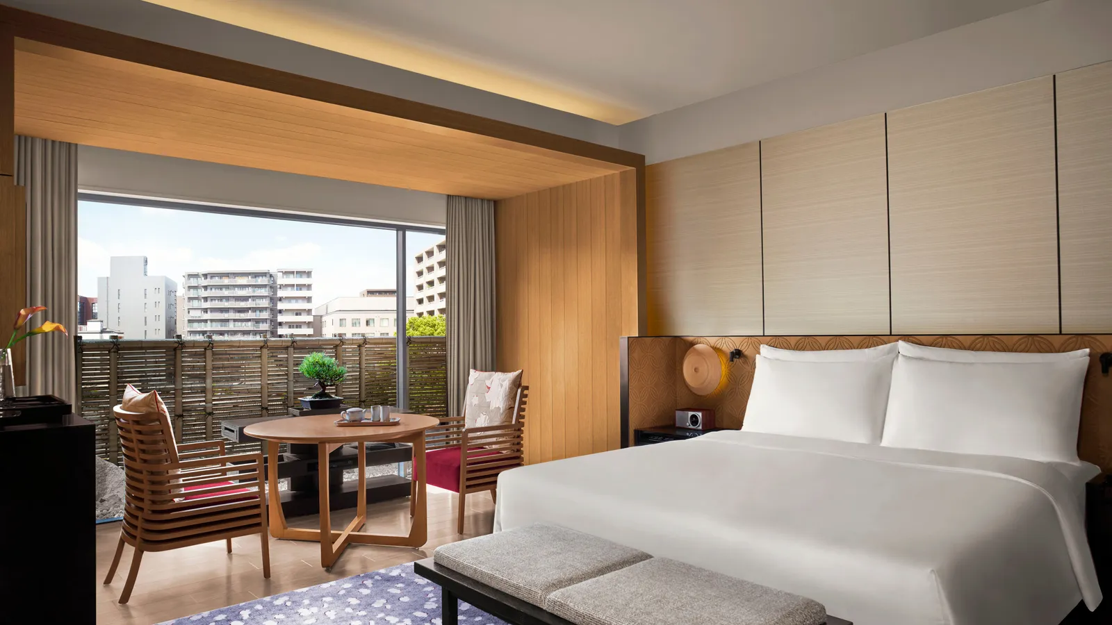 A Deluxe room with its king bed, a round table, 2 armchairs and the window onto the city’s buildings beyond a wooden screen.