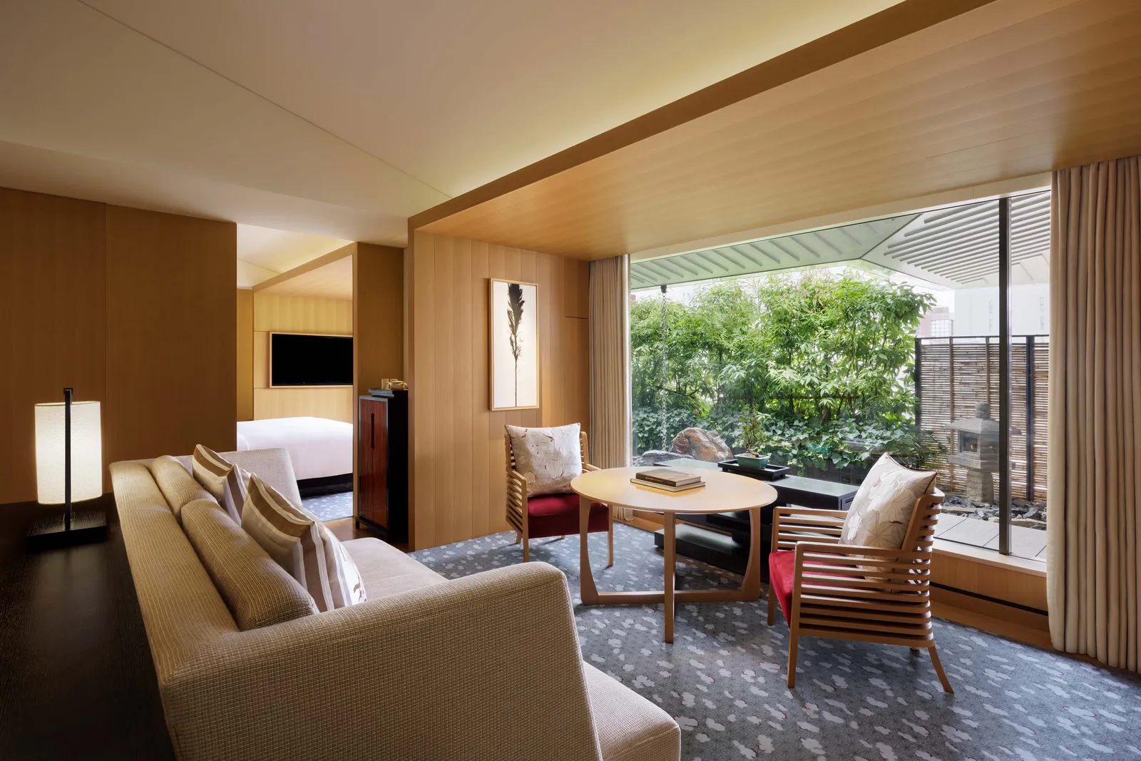 A Garden Suite’s living room: sofa, armchairs and a round table before the glass wall onto its private garden, the bedroom behind.