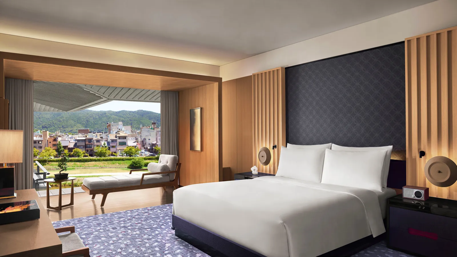 The Suite KAMOGAWA bedroom with its king bed, a chaise longue under the window and the river and hills beyond the terrace.
