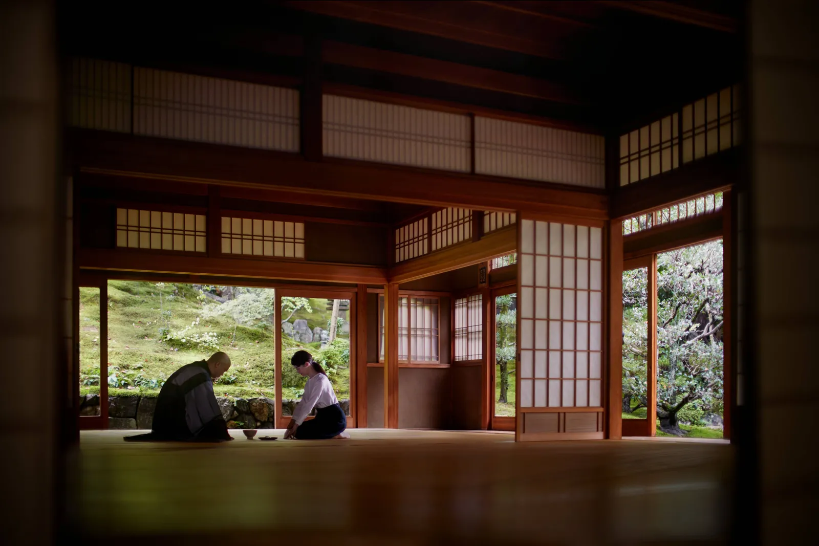 A monk and a guest kneeling on the wooden floor of a traditional hall that opens onto a moss garden.