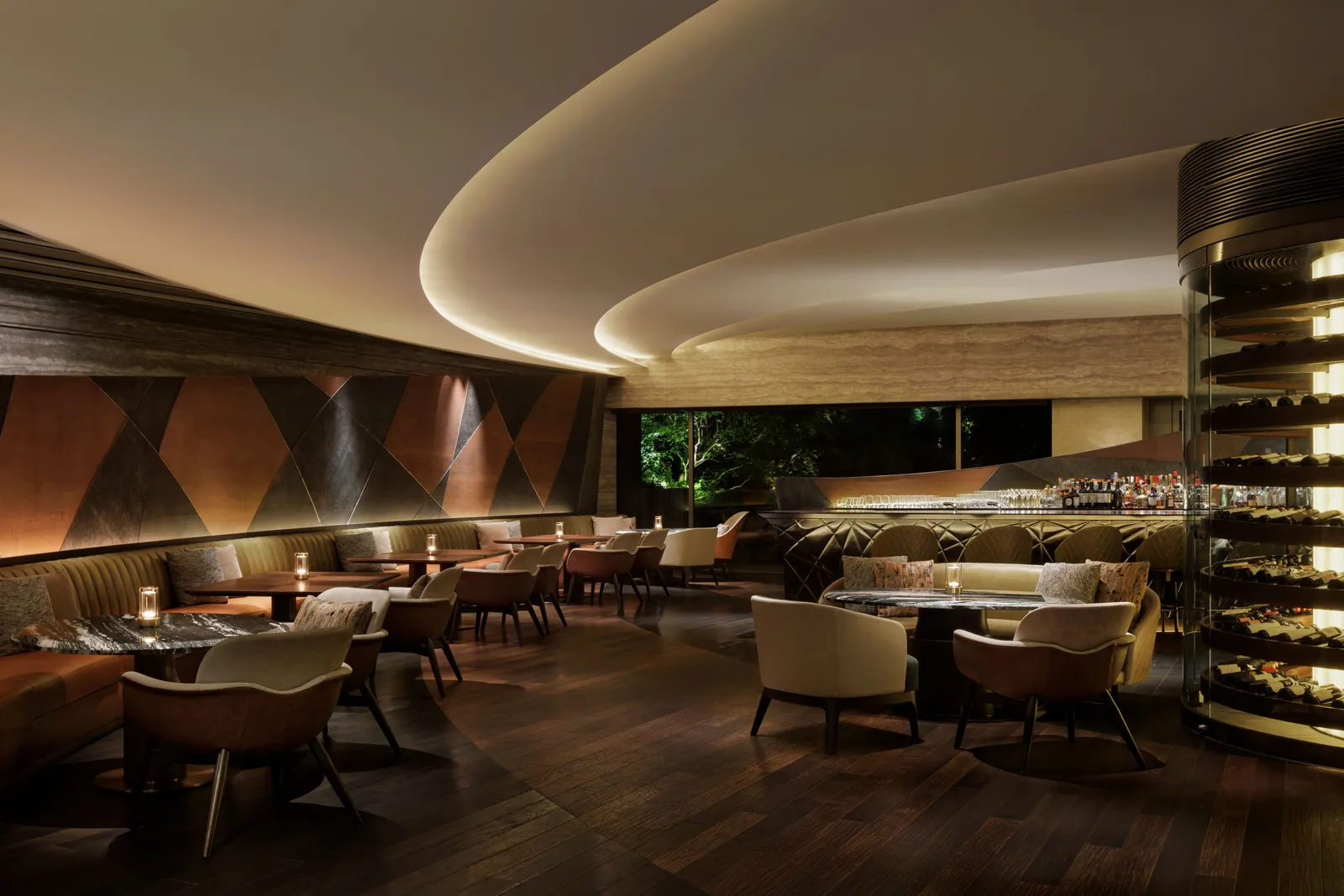 The Bar in the evening: low armchairs round marble tables, a curved lit ceiling and a tower of bottles on the right.