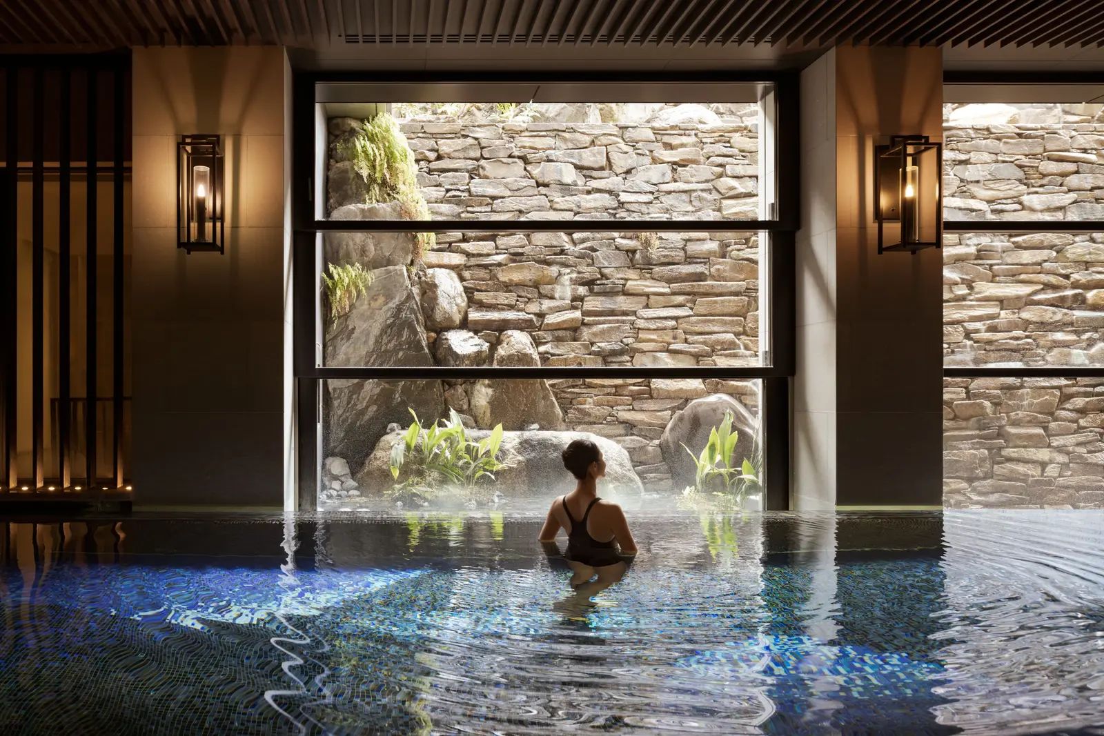 A woman in the indoor pool looking out at the dry-stone wall and the plants beyond the tall window.