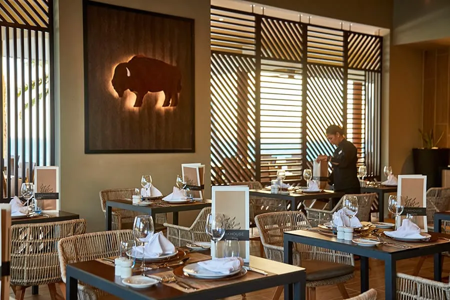 The Beef Steak House dining room with its laid tables, rattan chairs and a waiter at the back.
