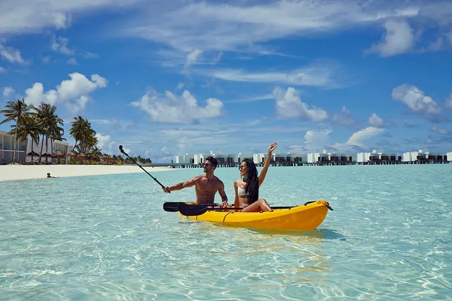 A couple in a yellow kayak on the lagoon, with the beach and palms behind.