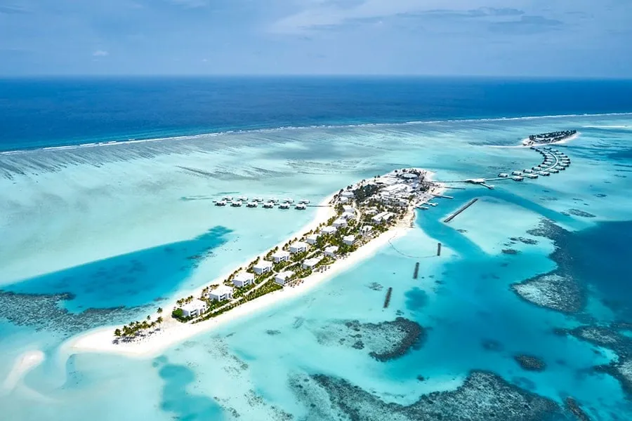 Kedhigandu from above: the strip of white sand with the buildings among the palms, the jetty of overwater suites and the reef around it.