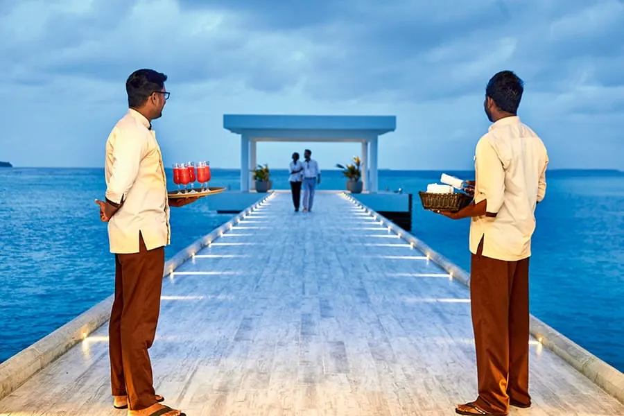 2 members of staff wait on the jetty with welcome drinks and towels as a couple walk in from the sea.