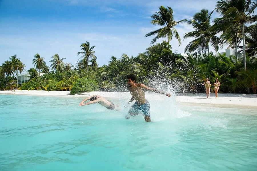 2 guests run and dive into the shallow lagoon in front of the beach, in a burst of spray, with the palms behind.