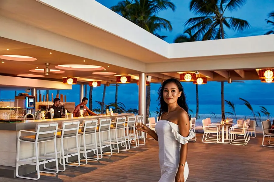 The Paradise bar at dusk: the long white counter with its stools, 2 bartenders and a young woman in a white dress against the sea.