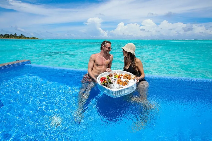A couple breakfast sitting in the private pool of an overwater suite, with a floating tray and the lagoon behind them.