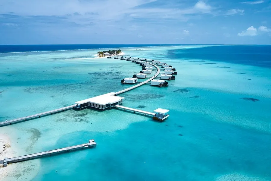 From above, the jetty of overwater suites curving through the lagoon towards the island, with the pavilion at the junction.