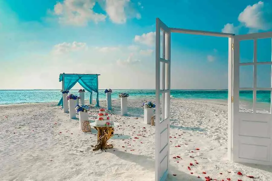 A beach wedding set-up: a white door standing open, petals on the sand, flowers and an arch draped in pale blue.