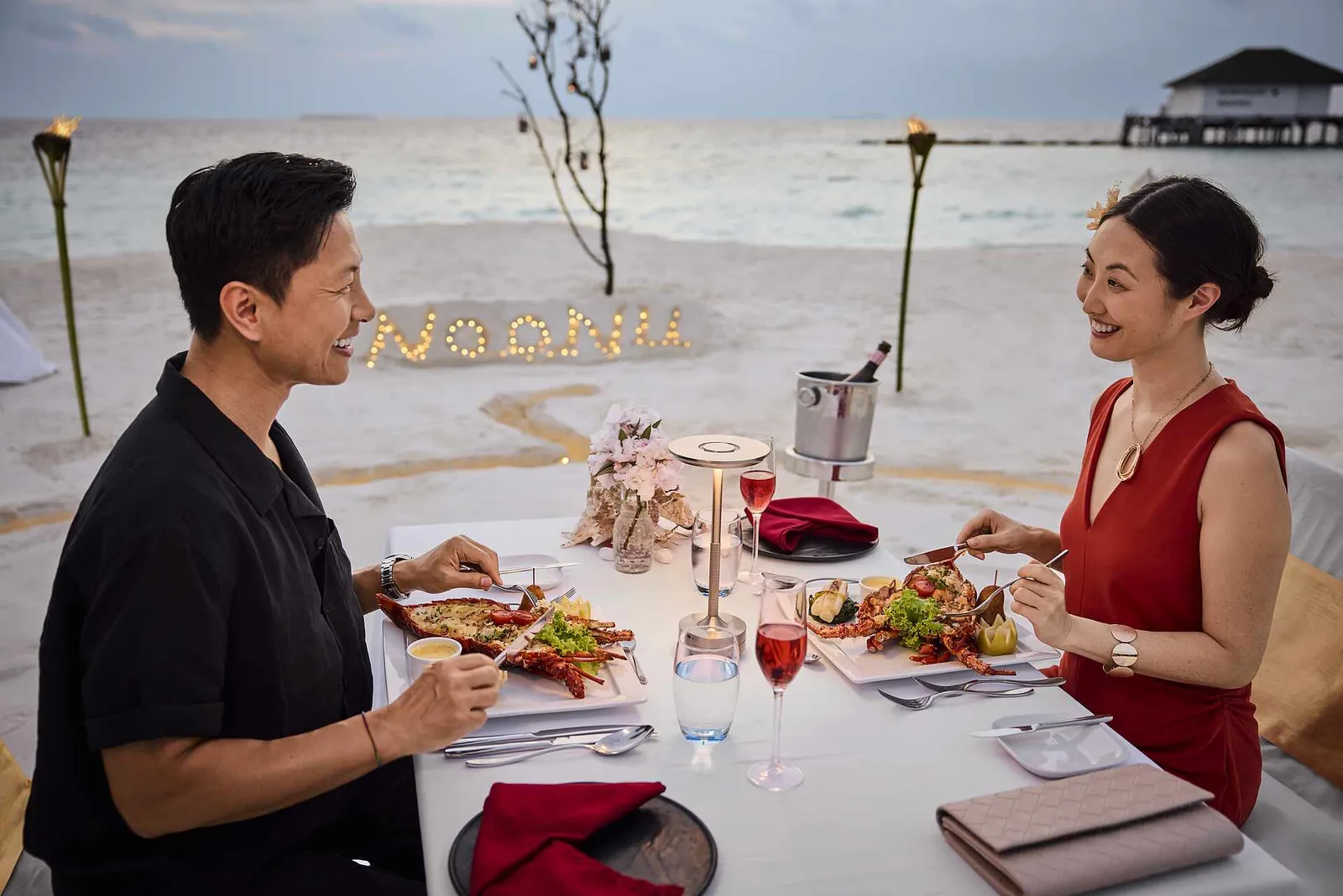 A couple’s dinner on the sand at sunset, with lobster, lit torches and the word Noonu spelt out in candles.